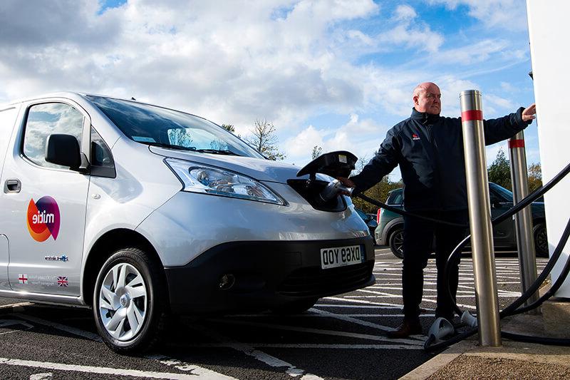 A Mitie branded electric van being charged, with a Mitie employee operating the charge point system at the front of the van